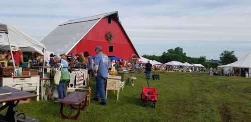 A vibrant outdoor market with vendors and shoppers near a red barn, surrounded by tents and greenery.