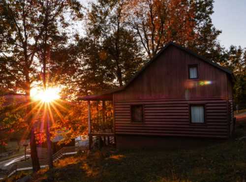 A rustic cabin surrounded by trees, with a vibrant sunset casting rays of light through the autumn foliage.