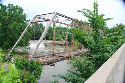 Rusty metal bridge over a river, surrounded by lush green trees and a concrete barrier in the foreground.