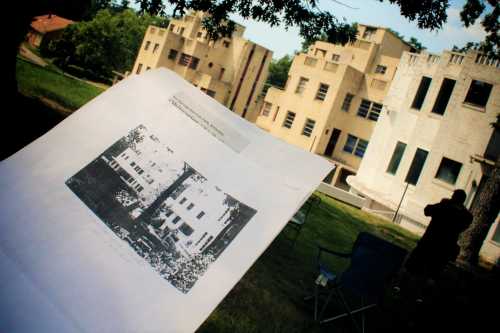 A printed architectural plan in the foreground, with modernist buildings visible in the background under a tree.