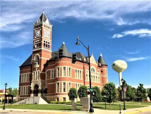 Historic brick courthouse with a clock tower, surrounded by trees and a water tower under a blue sky.