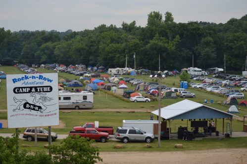 Aerial view of a campground with numerous tents and RVs, featuring a sign for Rock-n-Row Adventures Camp & Tube.