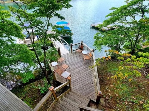 A wooden deck with chairs overlooks a serene lake surrounded by trees and boats docked nearby.