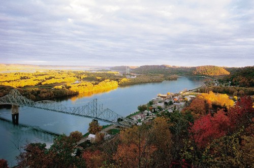 A scenic view of a river winding through autumn-colored trees, with a bridge and small town visible along the shore.