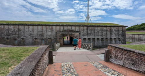 Two people walk towards a historic fort entrance under a blue sky with scattered clouds. Green grass surrounds the area.
