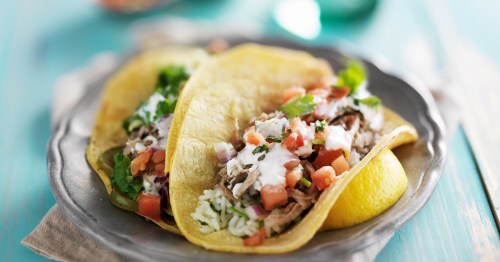 Two tacos filled with shredded chicken, rice, tomatoes, and greens, served on a silver plate with a colorful background.