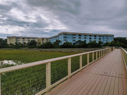 A wooden boardwalk leads to a blue building surrounded by green marshland under a cloudy sky.