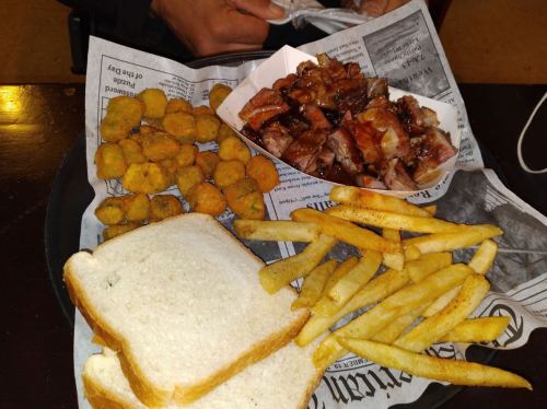 A plate of food featuring fried okra, barbecue meat, fries, and slices of white bread on newspaper.