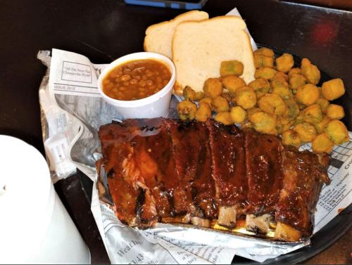 A plate of ribs with barbecue sauce, sides of baked beans, fried okra, and two slices of bread on a tray.