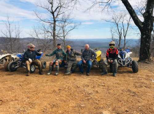 Five people sit on a dirt path with ATVs, smiling against a scenic backdrop of trees and hills.