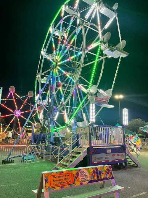 A brightly lit Ferris wheel at night, surrounded by colorful carnival lights and a sign for foot recovery.