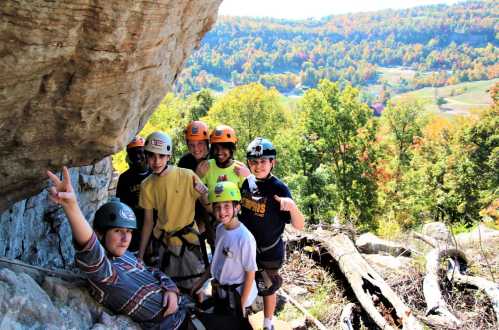 A group of seven kids in helmets poses for a photo outdoors, surrounded by trees and a scenic landscape.