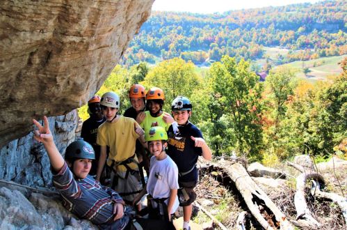 A group of seven kids in helmets poses for a photo outdoors, surrounded by trees and a scenic landscape.