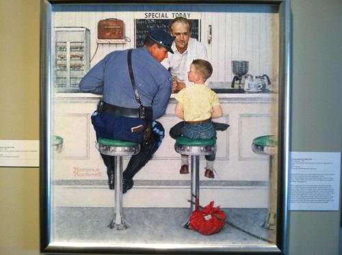 A police officer chats with a young boy at a diner counter, while a man behind the counter observes.
