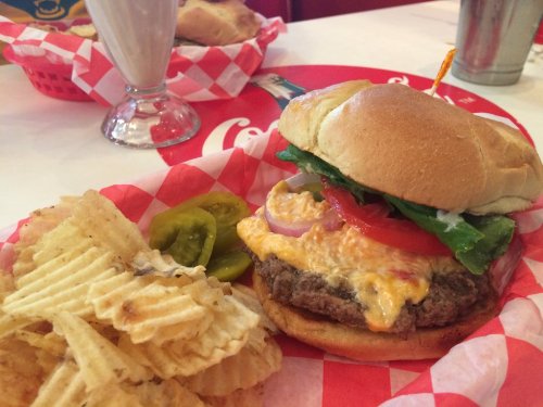 A juicy cheeseburger with lettuce, tomato, and pickles, served with a side of ridged potato chips on a red checkered plate.
