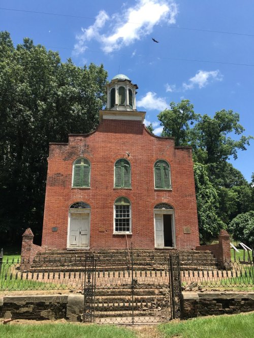 Historic red brick building with green shutters, a dome, and a fenced yard, surrounded by trees under a blue sky.