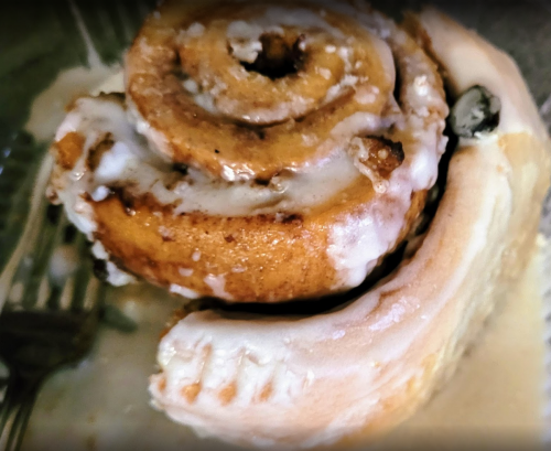 A close-up of a cinnamon roll drizzled with icing, partially on a plate with a fork beside it.