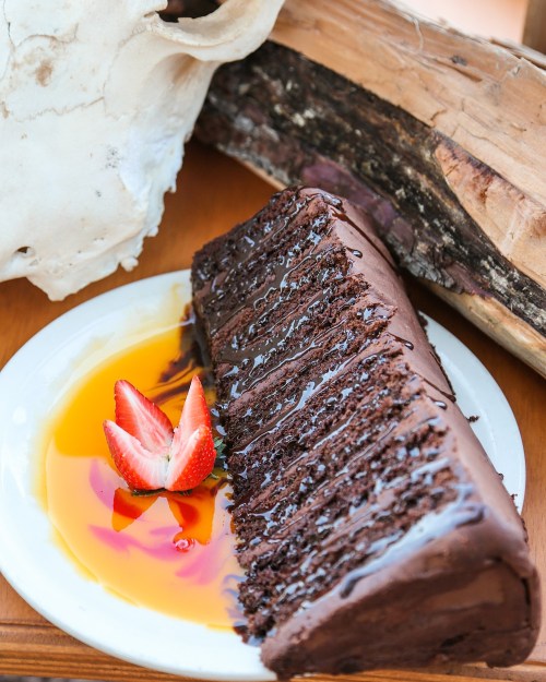A slice of chocolate cake drizzled with syrup, garnished with strawberries, next to a skull and wooden log.