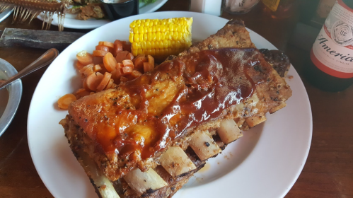 A plate featuring BBQ ribs with sauce, corn on the cob, and carrots, served with a drink in the background.