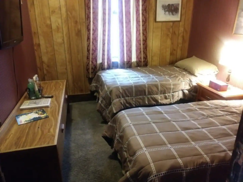 Cozy bedroom with two twin beds, wooden walls, a dresser, and a window with patterned curtains.