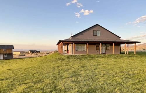 A modern house with a porch sits on a grassy hill, surrounded by open fields and a clear sky.