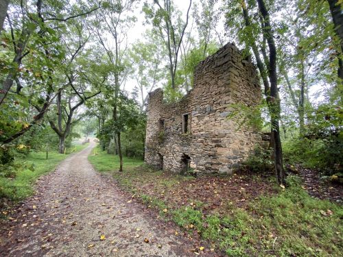 A stone ruin stands beside a gravel path, surrounded by trees and greenery on a cloudy day.