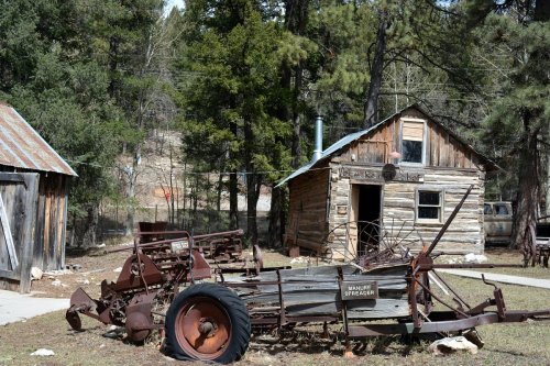 An old wooden cabin surrounded by vintage farming equipment in a forested area.