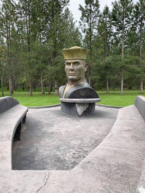 A large bust of a soldier in a park, surrounded by trees and a curved stone bench.
