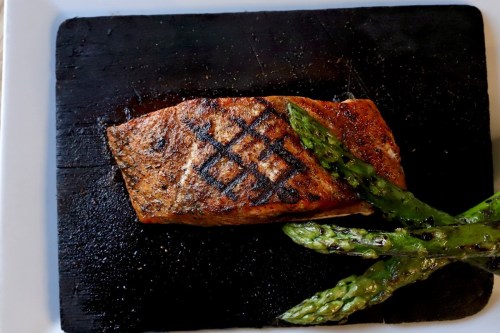 Grilled salmon fillet with a charred pattern, served on a black plate alongside grilled asparagus.