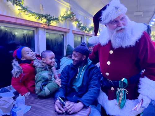 A joyful scene with Santa and two children, while a man sits at a table, all surrounded by festive decorations.