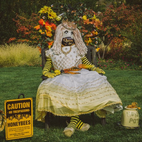 A whimsical scarecrow dressed as a queen, surrounded by flowers and a caution sign about honeybees.