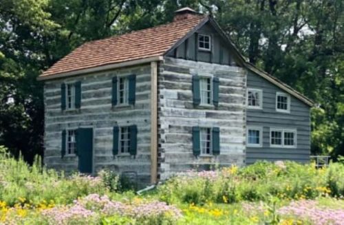 A rustic two-story house with a mix of log and wooden siding, surrounded by wildflowers and greenery.