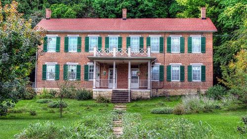A two-story brick house with green shutters, a front porch, and a landscaped yard surrounded by trees.