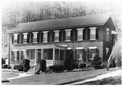 A historic two-story brick house with white shutters, a porch, and a staircase, surrounded by greenery.
