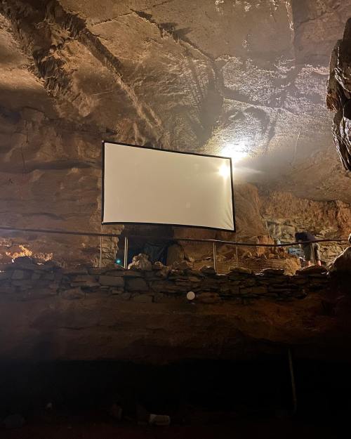A large blank screen is set up in a cave, illuminated by soft lighting from above.