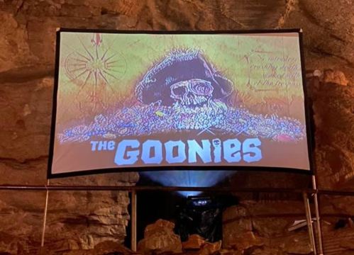 A projection of "The Goonies" on a screen in a cave, featuring a colorful skull and treasure illustration.