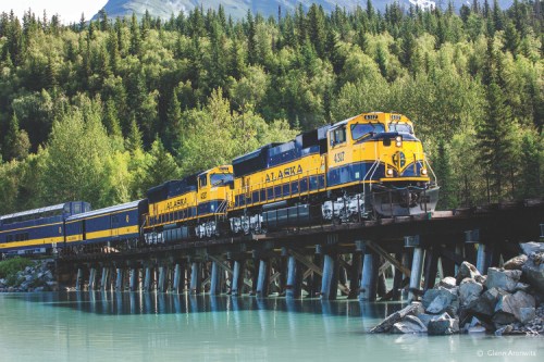 A yellow and blue Alaska Railroad train crosses a wooden bridge over a turquoise river, surrounded by lush greenery.