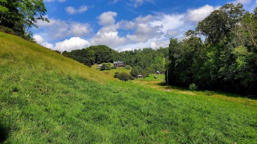 A lush green hillside with scattered trees and a few houses under a partly cloudy blue sky.