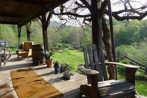 A cozy porch with wooden chairs, a rug, and plants, overlooking a lush green landscape and trees in the background.