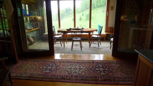 Cozy dining area with a wooden table and chairs, large windows overlooking a green hillside, and a patterned rug.