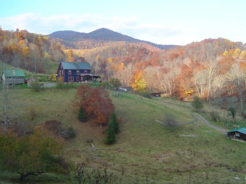 A scenic view of a blue house on a hillside, surrounded by autumn trees and mountains in the background.