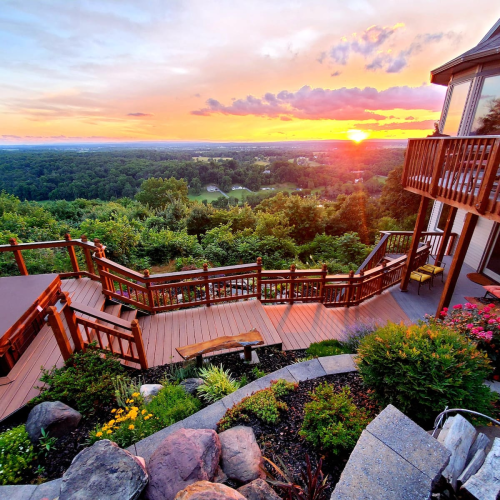 A scenic view from a deck overlooking lush greenery and a sunset in the background. Colorful flowers and rocks in the foreground.