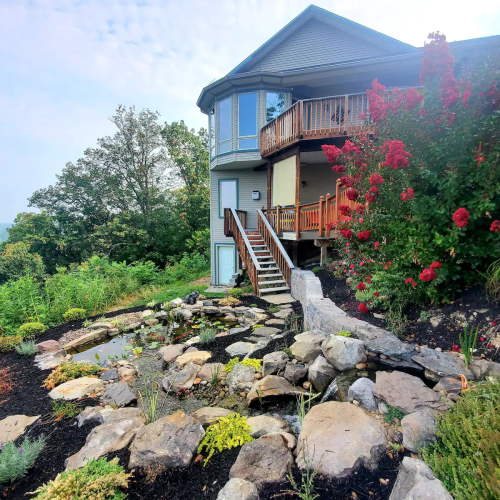 A modern house with a wooden deck, surrounded by lush greenery and a landscaped garden featuring rocks and plants.