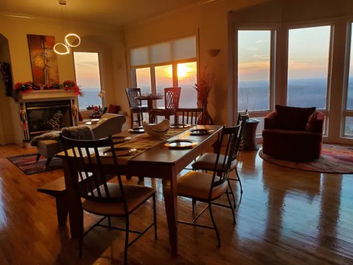 Cozy dining area with a wooden table, chairs, and a sunset view through large windows. Warm, inviting atmosphere.