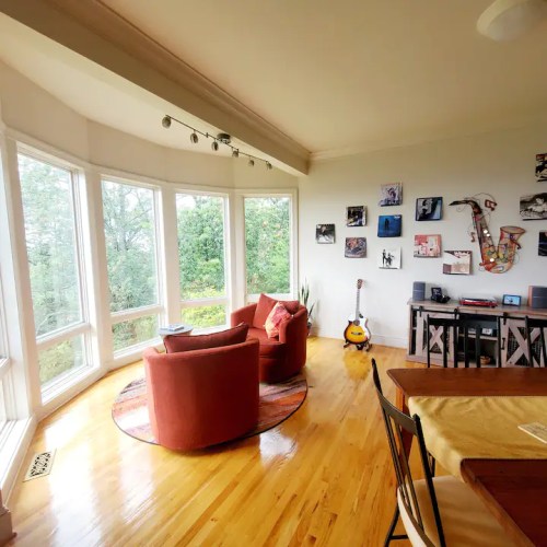 Cozy living room with large windows, two chairs, a guitar, and wall art, surrounded by greenery outside.