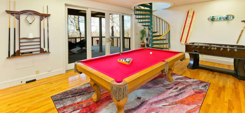 A game room featuring a red pool table, foosball table, and a spiral staircase, with large windows overlooking nature.