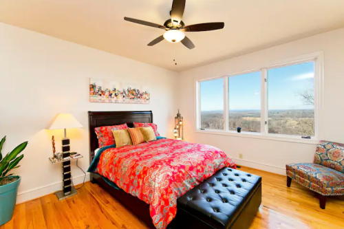 Bright bedroom with a colorful bedspread, large windows, and a cozy seating area, featuring wooden floors and a ceiling fan.