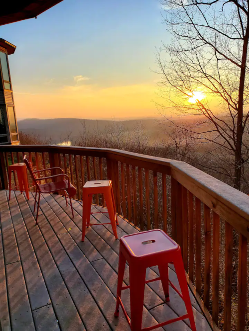 A wooden deck with orange stools overlooks a sunset over a valley, surrounded by trees.