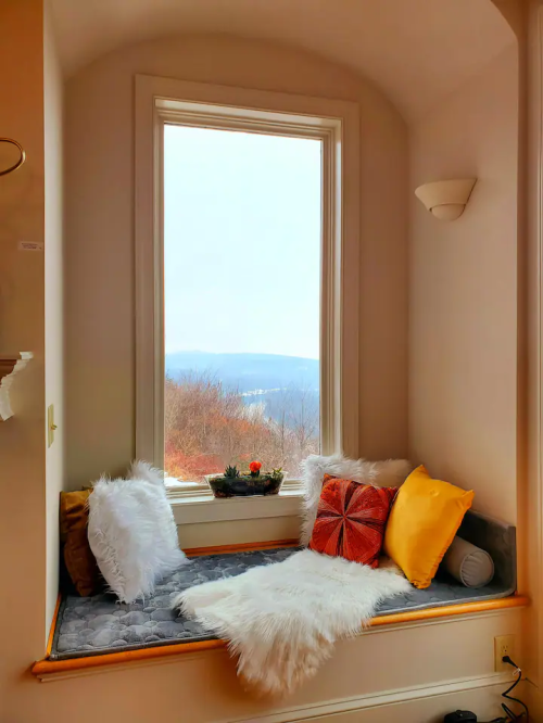 Cozy window nook with cushions and a view of mountains, featuring a small plant and decorative pillows.