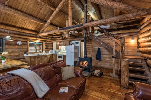 Cozy log cabin interior with wooden beams, a fireplace, and a staircase, featuring a living area and kitchen.
